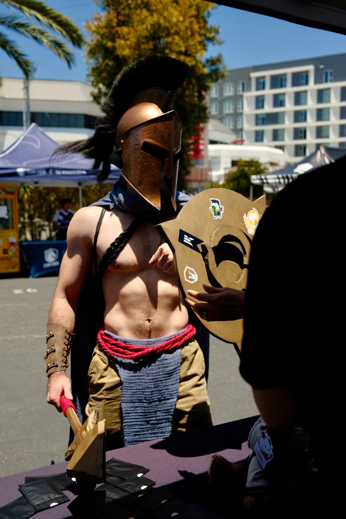 A cosplayer in a Spartan helmet holding a sticker-covered shield at an event