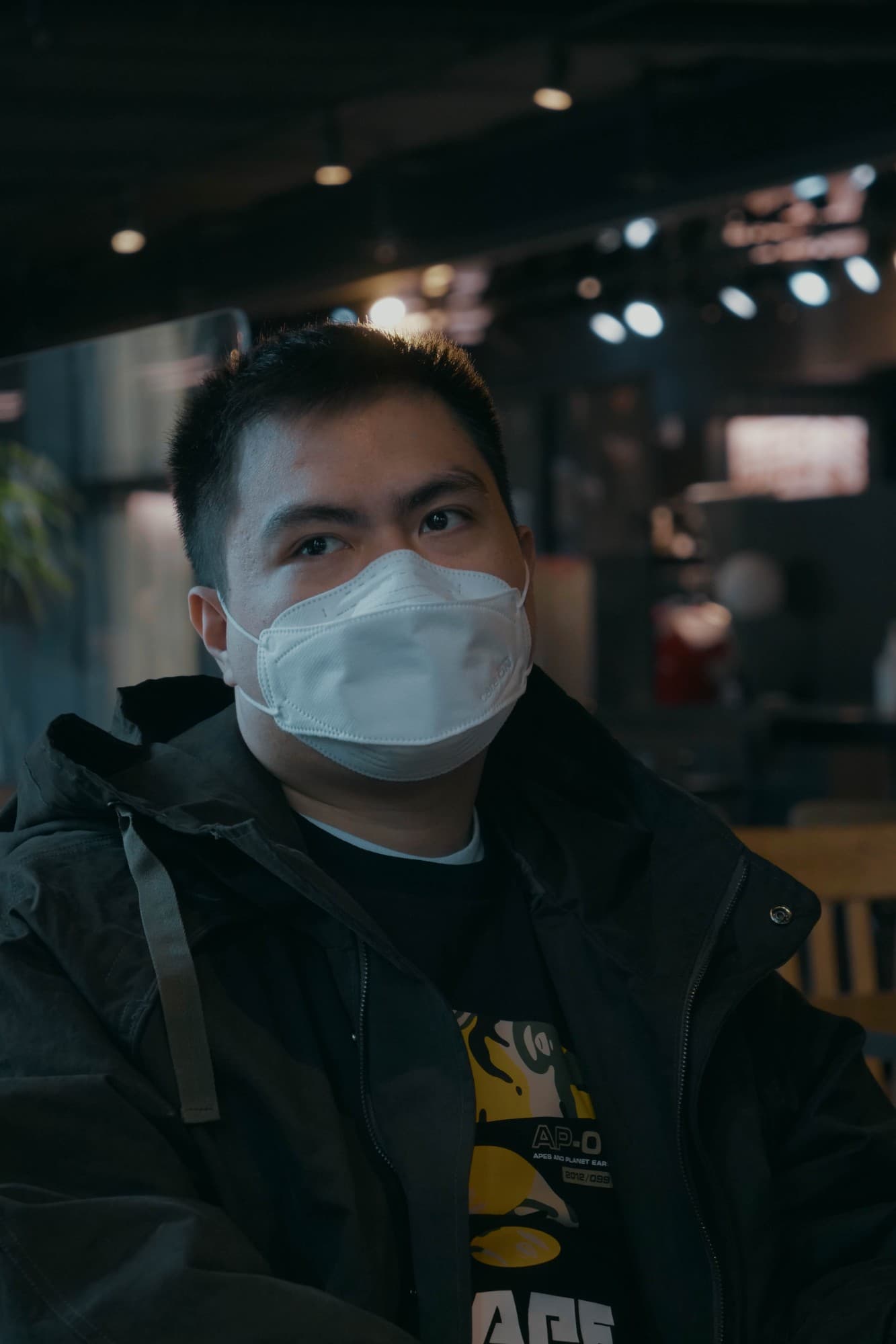 A man in a white mask and black jacket sitting in a dimly lit cafe